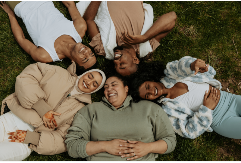 Social Rest Image. Image of 5 people from diverse backgrounds with diverese bodies laying on the ground with their heads all touching laughing and smiling.